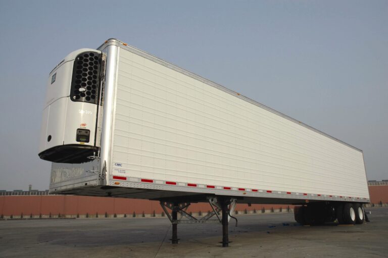 A large, white refrigerated semi-trailer is parked on a paved lot under a clear sky, with its cooling unit visible at the front and no truck cab attached.