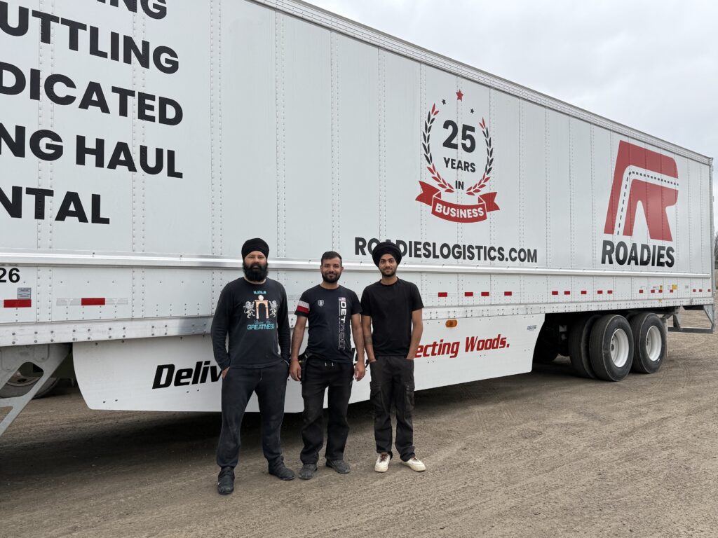 Three men stand in front of a white Roadies Logistics trailer that displays "25 Years in Business" and company branding. The trailer is parked on a gravel surface under a cloudy sky.