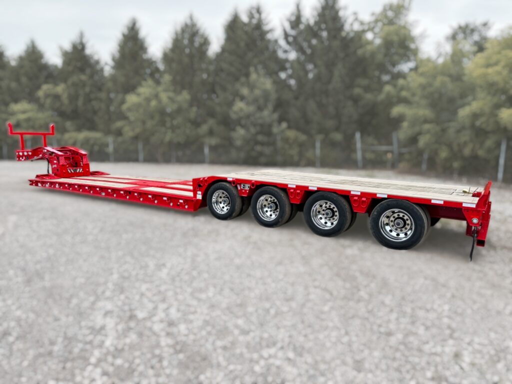 A bright red, multi-axle flatbed trailer with four wheels on each side is parked on a gravel surface, with trees and a cloudy sky in the background.
