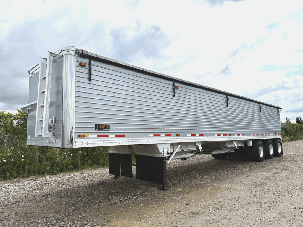 A large silver grain trailer with multiple axles is parked on a gravel surface under a cloudy sky, surrounded by green vegetation.