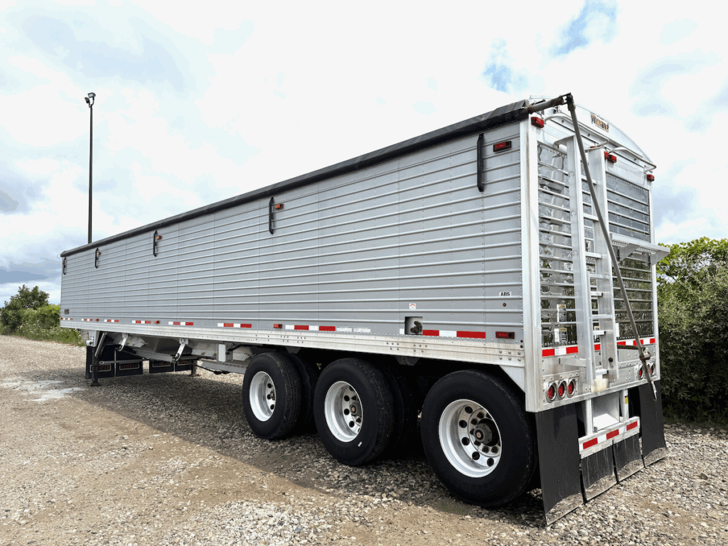 A large silver grain trailer with black wheels is parked on a gravel lot under a partly cloudy sky, with greenery visible in the background.